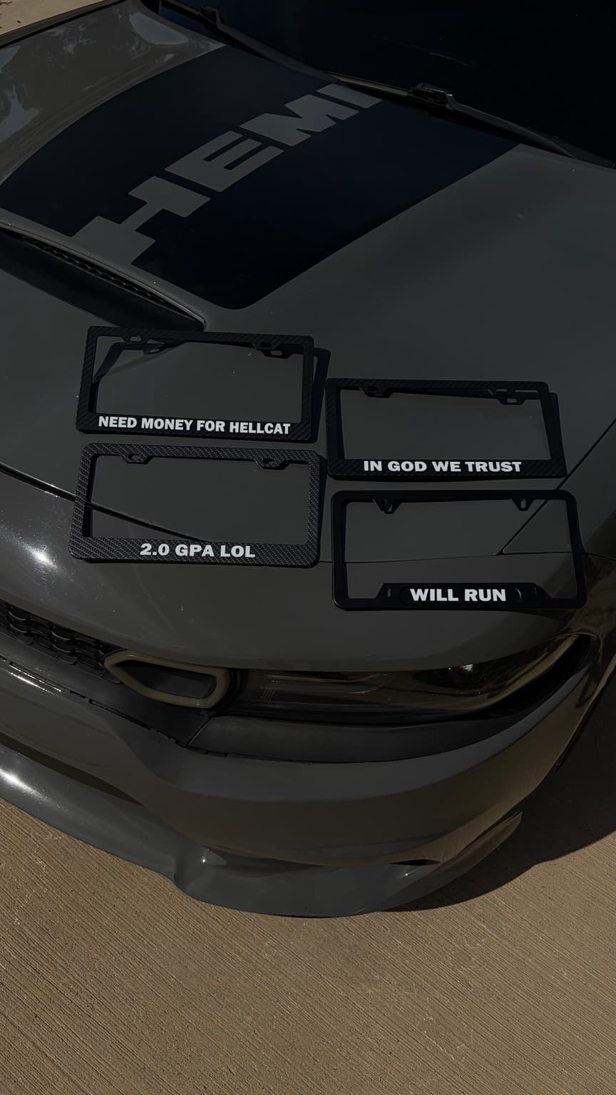 Four custom license plate frames in black aluminum and carbon fiber, with white bottom text, on a gray Charger hood